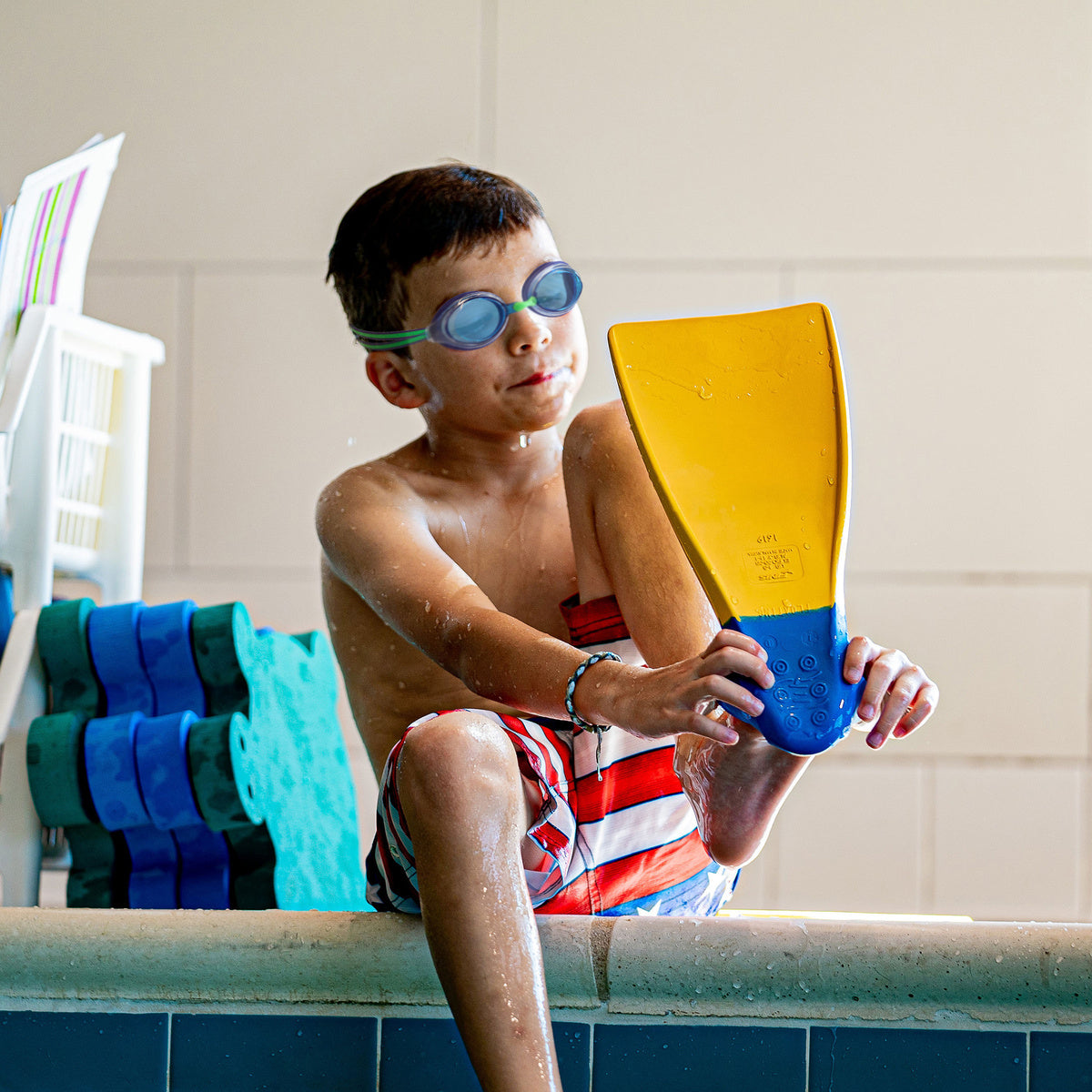 Alt Text: Boy at poolside adjusting swim fin while wearing FINIS Racer Ripple goggles