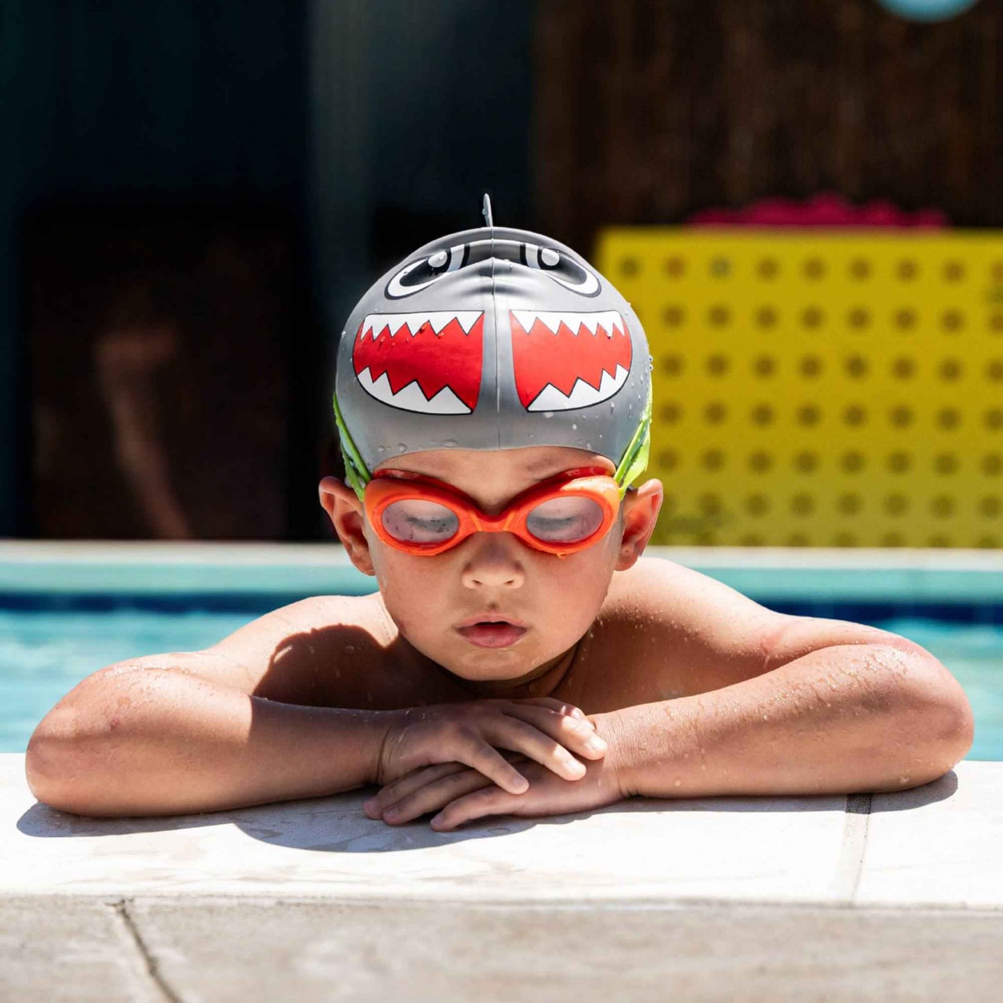 Child wearing FINIS Orange scented swim goggles resting at the pool edge in shark cap